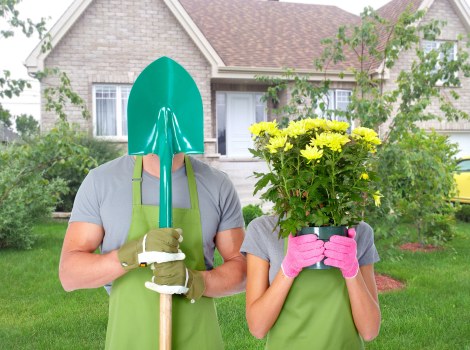 A gardener and a woman are standing in front of a residential house with a well-maintained lawn and garden. The gardener is wearing a grey t-shirt, green gardening gloves, and a green apron, holding a bright blue shovel with the blade facing upward. The woman is dressed in a grey top and pink gardening gloves, holding a large pink pot filled with yellow daisy-like flowers, partially obscuring her face. Behind them, the front garden features neatly trimmed grass, various small shrubs, and young trees with green foliage. The house has light-colored brick walls, white window frames, and a small porch, with a clear sky overhead indicating daytime. The scene depicts outdoor gardening activity suitable for landscaping and lawn care services within the Deptford area, demonstrating general garden maintenance that could be offered by Hedge Trimming Deptford. The natural setting and garden details support the context of professional outdoor garden and landscaping services in the local area.