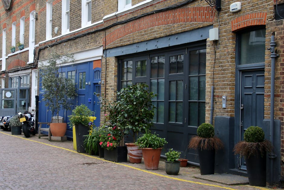 A row of small urban garden spaces along a cobblestone street in Deptford Dockyard, featuring a variety of potted plants including lush green shrubs, flowering plants with pink blossoms, and small top