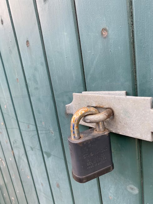 Close-up view of a black padlock securing a green wooden garden gate with vertical panels. The padlock is attached to a metal latch mounted on the gate, with the shackle showing signs of rust and corr